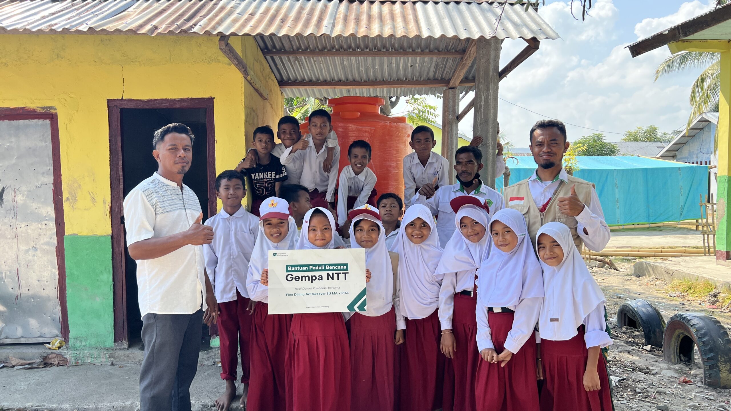 Para siswa MIS Mujahidin Arabi dan relawan berfoto bersama membawa papan bantuan sekolah darurat Manggarai Timur.