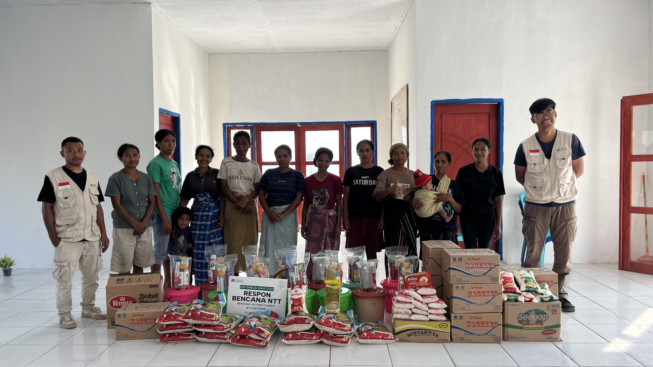 Tim Relawan Nusantara dan penerima manfaat berfoto bersama di balik tumpukan paket sembako, hygiene kit, dan air kemasan bantuan Second Chance by Evie Home.
